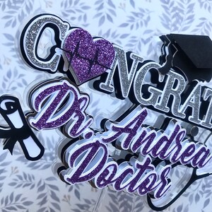 以下が含まれることがあります： 紫と銀のグリッターケーキトッパーに「おめでとうございます」と「Dr. Andrea Doctor」の文字が書かれています。トッパーには、卒業帽、心臓の鼓動のシンボルが描かれたハート、巻物も含まれています。