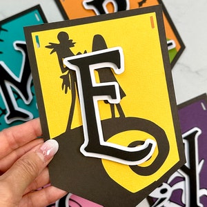 Puede incluir: Un banner de Halloween con las letras "MERRY" en negro con contornos blancos. Cada letra est&aacute; sobre un fondo de color diferente. La "M" est&aacute; sobre turquesa, la "E" est&aacute; sobre amarillo, la "R" est&aacute; sobre naranja y la "Y" est&aacute; sobre morado. Cada letra tiene una silueta de personaje de Halloween diferente.