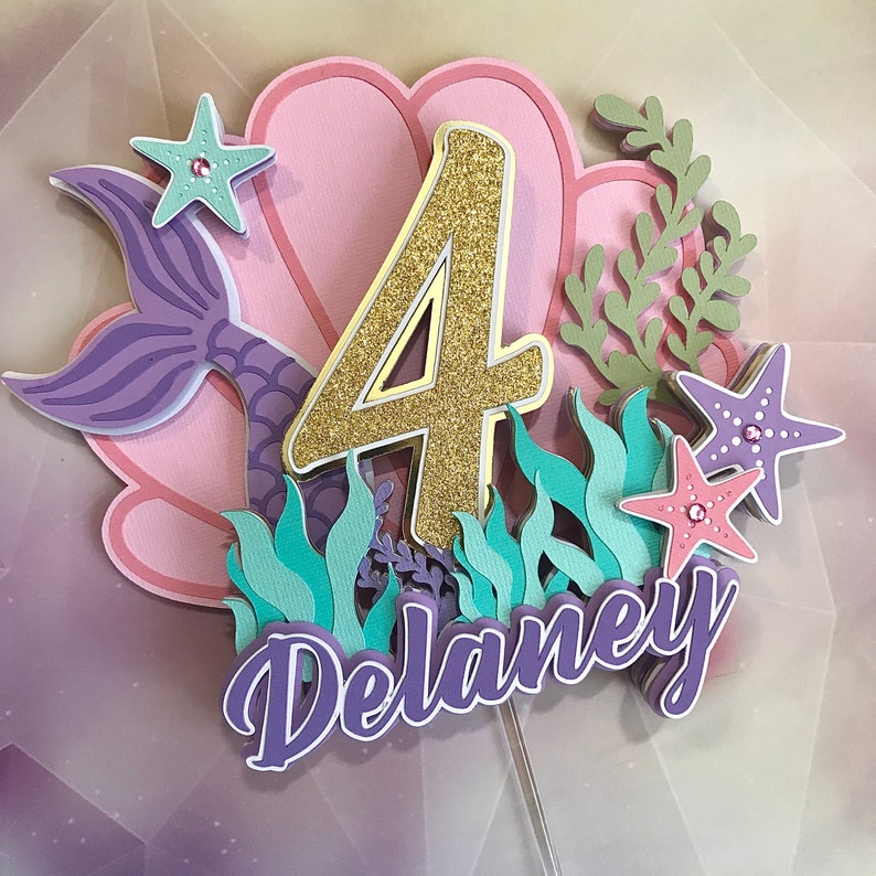 May include: A purple mermaid tail, a pink seashell, and a gold glitter number four are part of a cake topper with a turquoise and purple seaweed design. The name "Delaney" is written in purple on the cake topper.
