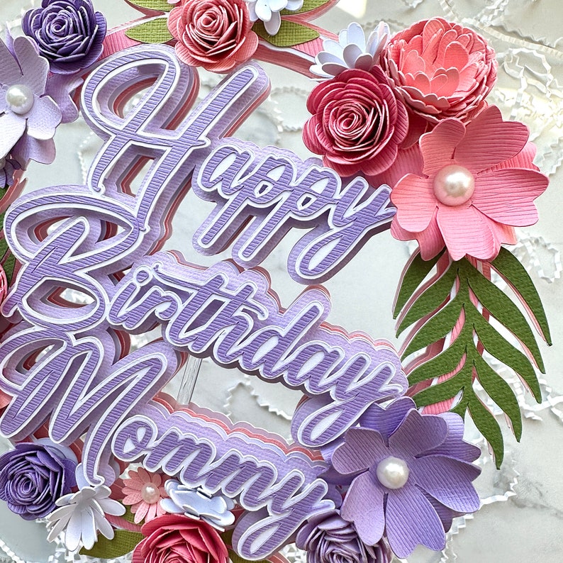 Op de afbeelding: Paarse en roze papieren bloemen versieren een paarse taarttopper met de woorden "Happy Birthday Mommy".