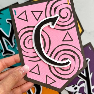 Puede incluir: Una pancarta de papel rosa y negro con la letra "C" en blanco y negro. La pancarta tiene un patr&oacute;n de espiral y tri&aacute;ngulos rosas.