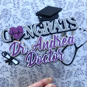 以下が含まれることがあります： 卒業帽、聴診器、卒業証書、そして「Congrats Dr. Andrea Doctor」の文字が書かれた、紫と銀のキラキラしたケーキトッパーです。