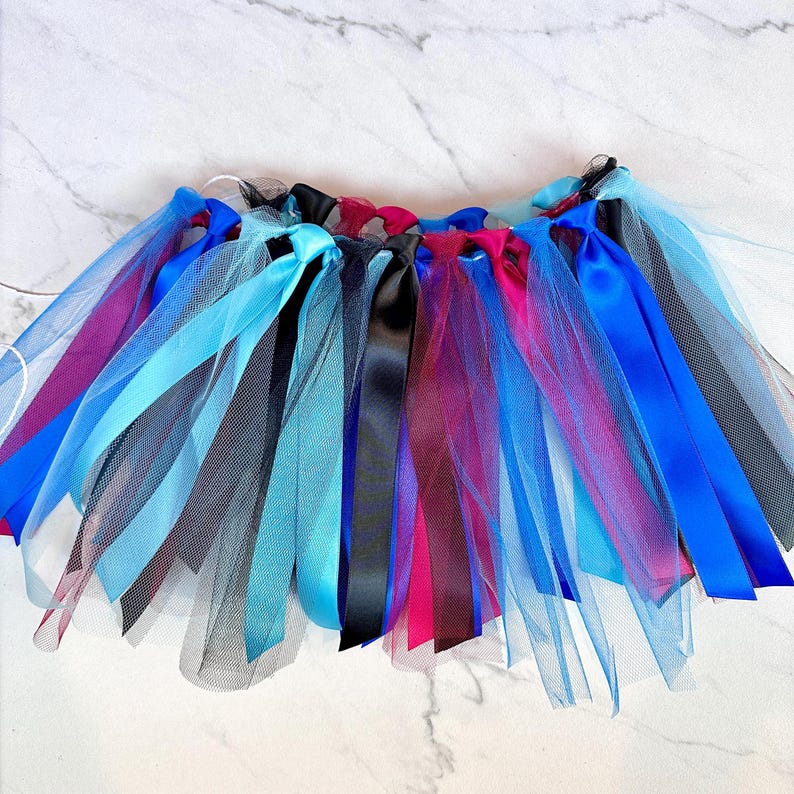 May include: A vibrant tutu skirt with layers of tulle and satin ribbons. The skirt features blue, black, pink, and purple ribbons. The tulle is light blue and black. Suitable for dance or costume purposes.