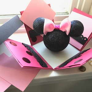 May include: A pink and black explosion box with a black glitter Minnie Mouse head in the center. The box is decorated with black polka dots and lace.