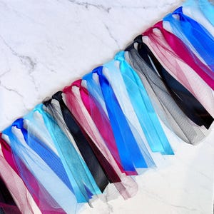 May include: A colourful tulle garland featuring ribbons in blue, pink, black, and white. The ribbons are attached to a mesh base, creating a decorative and festive appearance. Ideal for party decorations or craft projects.