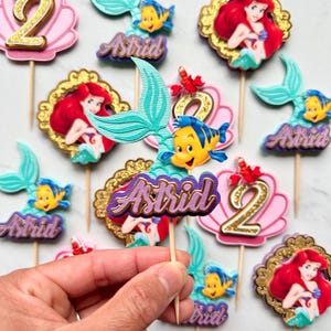 May include: A collection of decorative cupcake toppers featuring the name Astrid in purple script, alongside images of a mermaid, a yellow fish, a red crab, and the number 2. Each topper is attached to a wooden pick for placement in food.