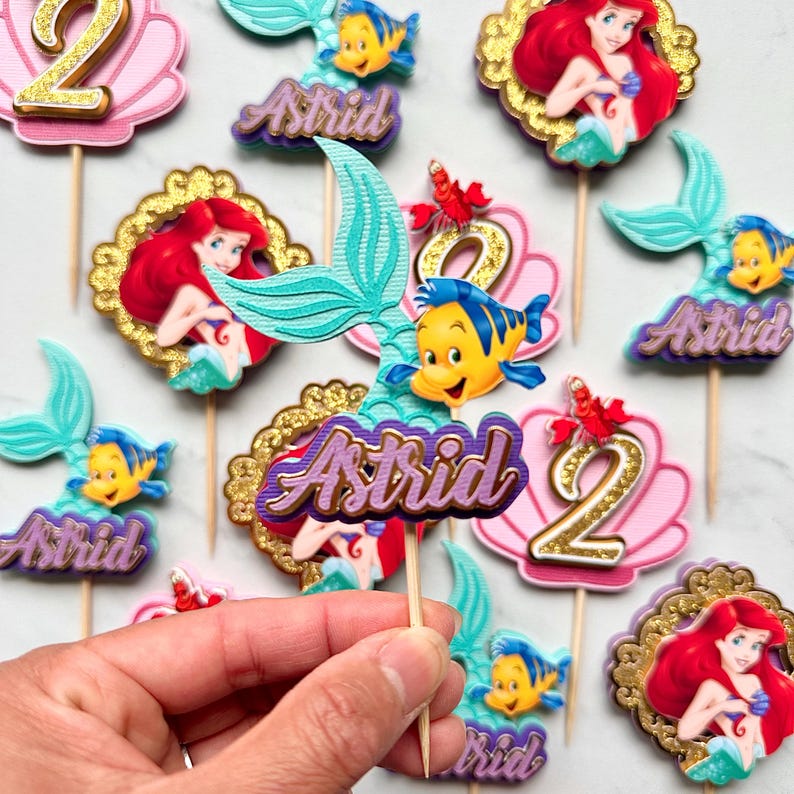 May include: A collection of decorative cupcake toppers featuring the name Astrid in purple script, alongside images of a mermaid, a yellow fish, a red crab, and the number 2. Each topper is attached to a wooden pick for placement in food.