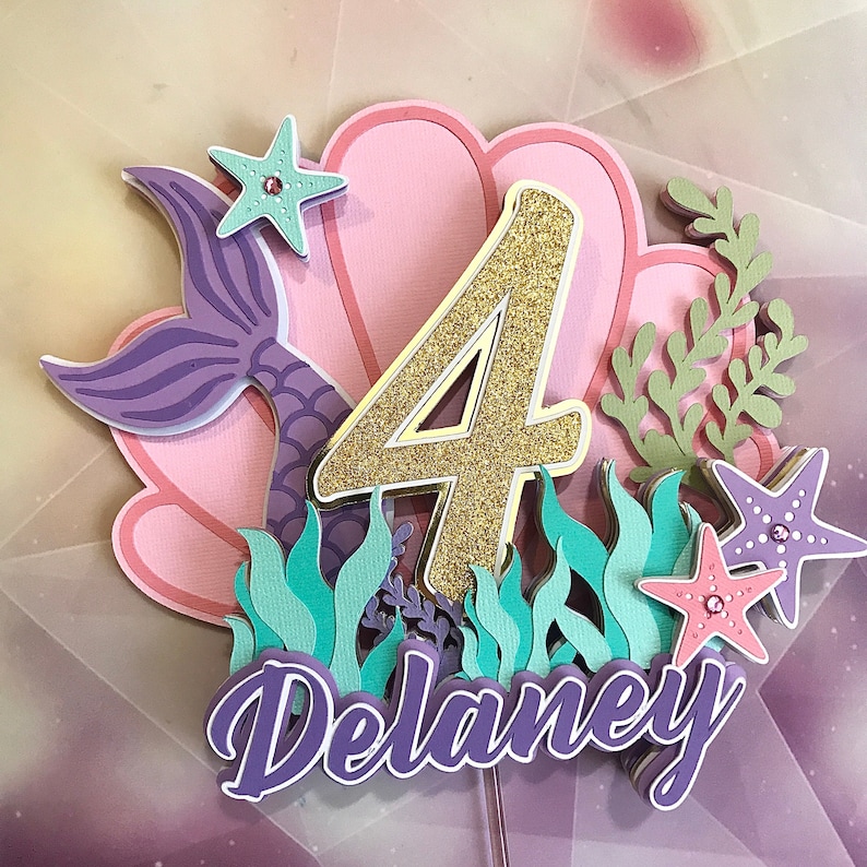 May include: A purple mermaid tail and a pink seashell with a gold glitter number 4 on top. The number is surrounded by green seaweed and pink starfish with glitter accents. The name Delaney is written in purple below the number.