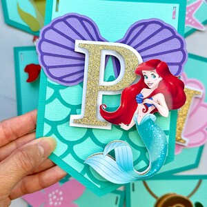 Puede incluir: Una pancarta con tem&aacute;tica de sirena, con la letra "P" en purpurina dorada, una concha morada y una ilustraci&oacute;n de sirena. Se ven otras pancartas con letras y decoraciones con tem&aacute;tica marina.
