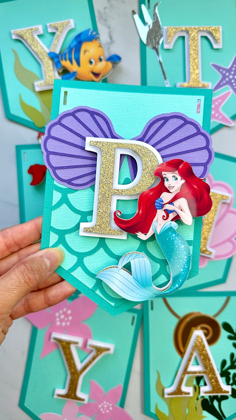Puede incluir: Una pancarta con tem&aacute;tica de sirena, con la letra "P" en purpurina dorada, una concha morada y una ilustraci&oacute;n de sirena. Se ven otras pancartas con letras y decoraciones con tem&aacute;tica marina.