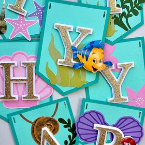 Puede incluir: Un cartel con banderines individuales que deletrean "HAPPY BIRTHDAY". Cada bander&iacute;n es de color turquesa con letras doradas con purpurina y adornos con tem&aacute;tica marina, incluyendo una estrella de mar, una concha y personajes de La Sirenita.