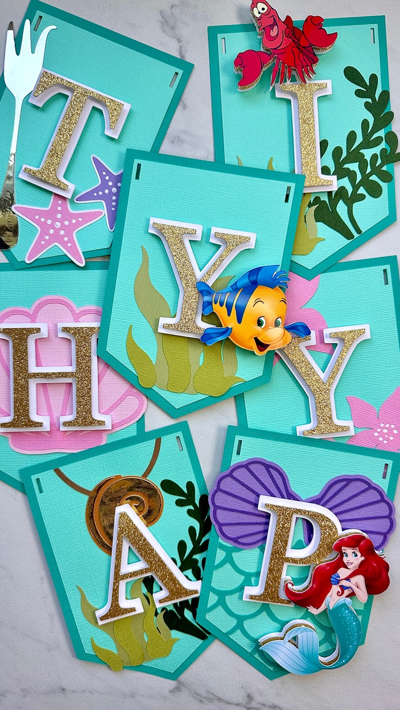 Puede incluir: Un cartel con banderines individuales que deletrean "HAPPY BIRTHDAY". Cada bander&iacute;n es de color turquesa con letras doradas con purpurina y adornos con tem&aacute;tica marina, incluyendo una estrella de mar, una concha y personajes de La Sirenita.