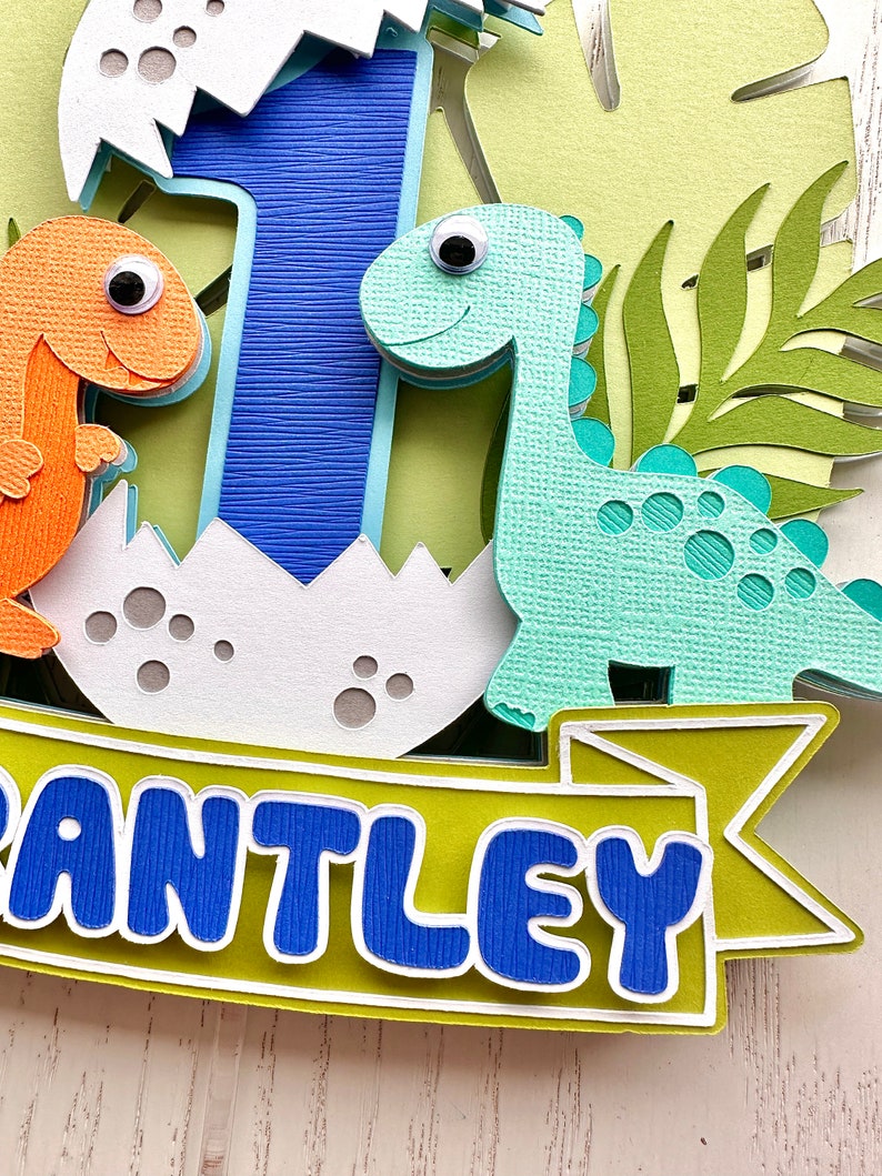 Pu&ograve; includere: Un topper per torta a tema dinosauro, di colore verde, blu e arancione, con il nome "ANTLEY" in lettere blu su uno stendardo verde. Il topper comprende un numero uno blu, un dinosauro verde e un dinosauro arancione.