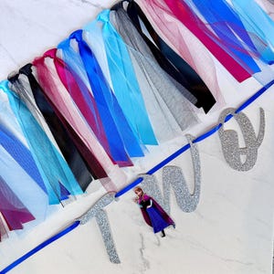 May include: A birthday banner spelling "TWO" in silver glitter. The banner is decorated with a cascade of tulle and ribbons in shades of blue, black, and burgundy. A small Anna figure is attached to the banner.