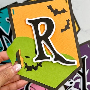 Puede incluir: Un banner de Halloween con un esquema de color verde, naranja y negro. El banner presenta la letra "R" en negro con un contorno blanco, un personaje verde de Jack Skellington y murci&eacute;lagos negros sobre un fondo naranja.