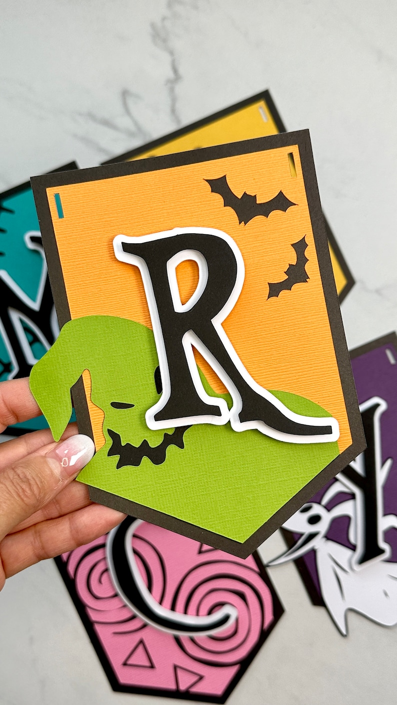 Puede incluir: Un banner de Halloween con un esquema de color verde, naranja y negro. El banner presenta la letra "R" en negro con un contorno blanco, un personaje verde de Jack Skellington y murci&eacute;lagos negros sobre un fondo naranja.