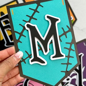 Puede incluir: Una pancarta azul turquesa con una letra negra "M" en un contorno blanco. La pancarta tiene un patr&oacute;n cosido alrededor de los bordes. La pancarta forma parte de una pancarta de Halloween.