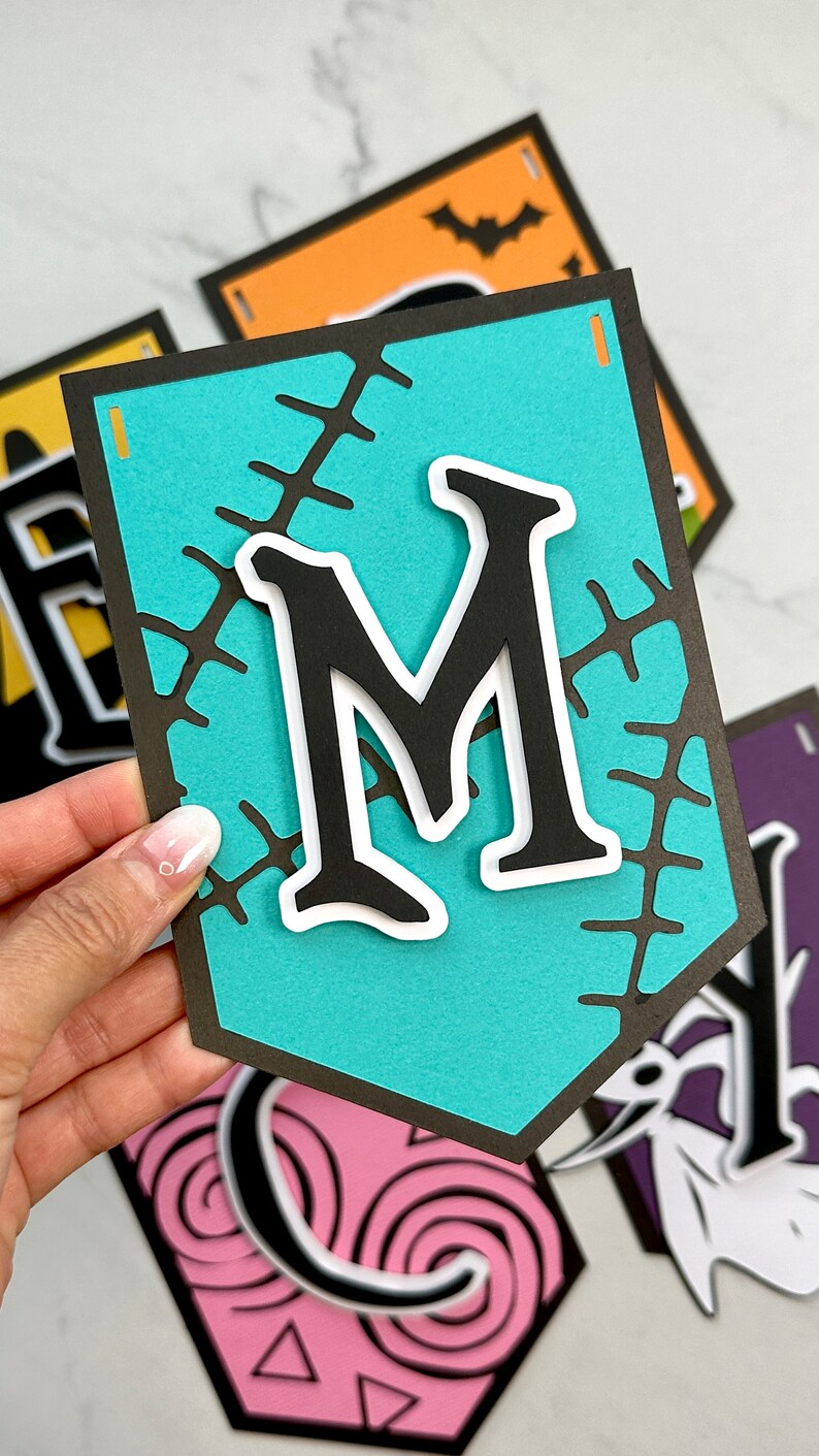 Puede incluir: Una pancarta azul turquesa con una letra negra "M" en un contorno blanco. La pancarta tiene un patr&oacute;n cosido alrededor de los bordes. La pancarta forma parte de una pancarta de Halloween.