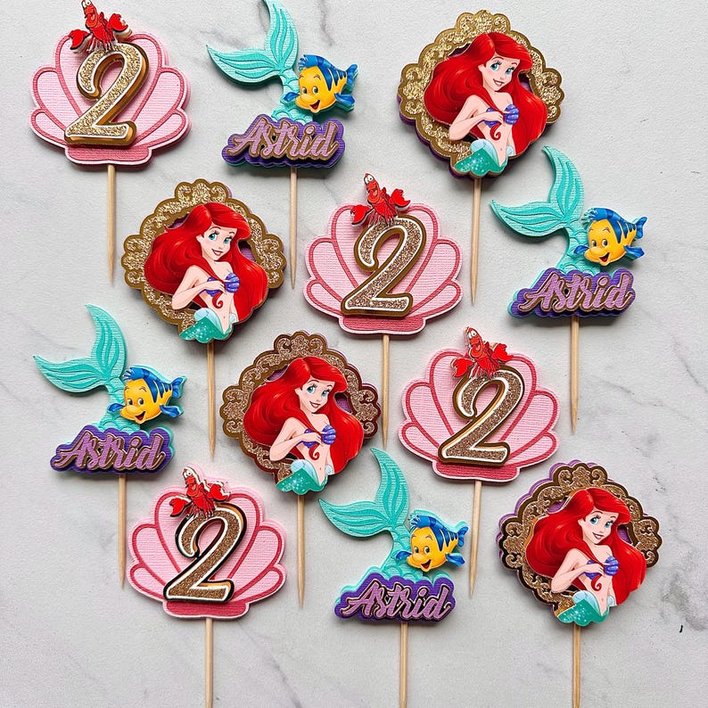 May include: A collection of cake toppers featuring The Little Mermaid characters, including Ariel, Flounder, and Sebastian. The set includes pink seashell cutouts with the number 2, mermaid tails with the name Astrid, and Ariel in gold frames.