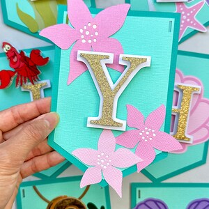 Puede incluir: Un banner con banderines turquesas con letras doradas con purpurina, flores rosas y recortes de personajes. El banner deletrea un nombre o frase, con personajes como Flounder, Sebasti&aacute;n y Ariel.