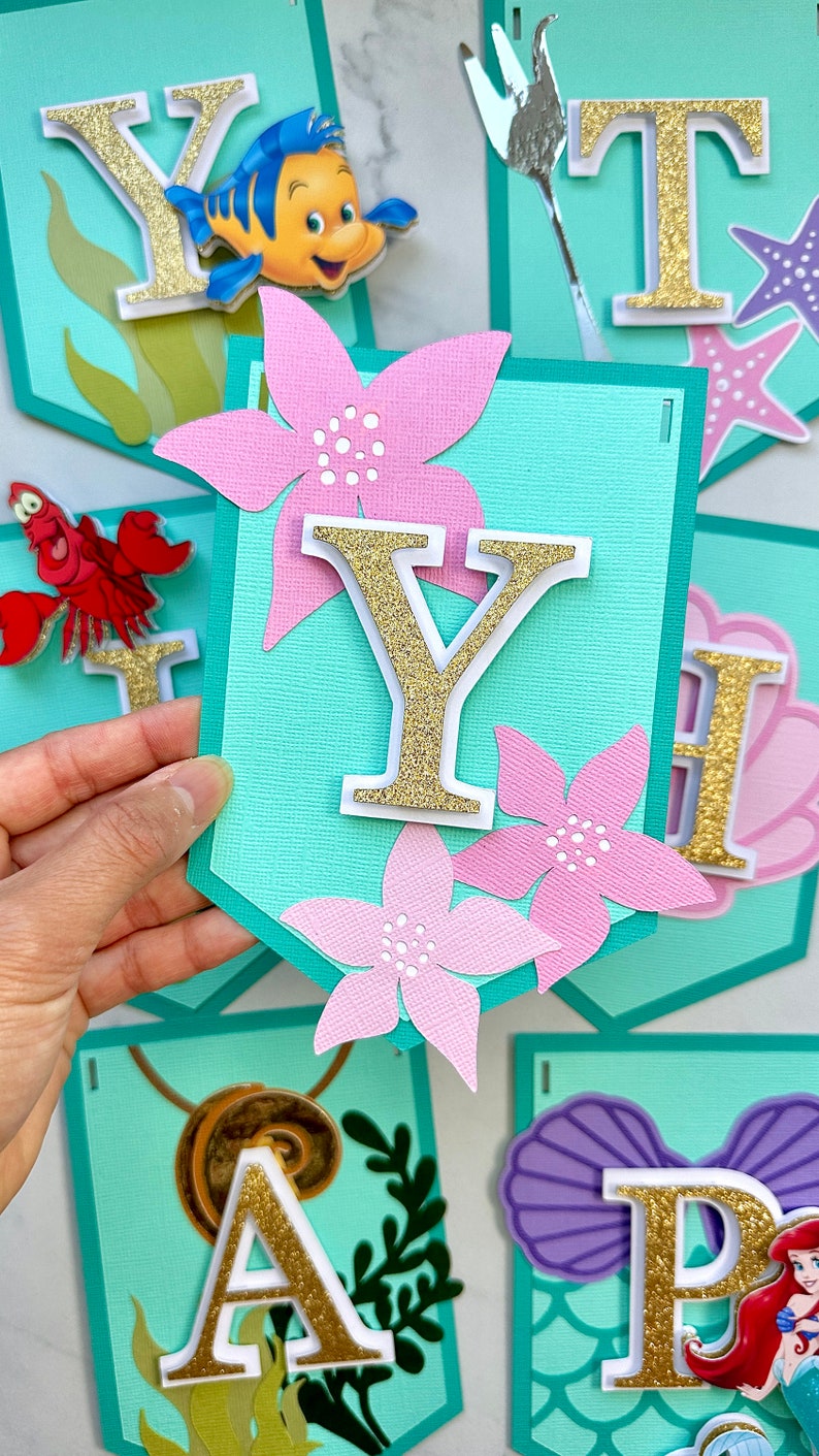 Puede incluir: Un banner con banderines turquesas con letras doradas con purpurina, flores rosas y recortes de personajes. El banner deletrea un nombre o frase, con personajes como Flounder, Sebasti&aacute;n y Ariel.