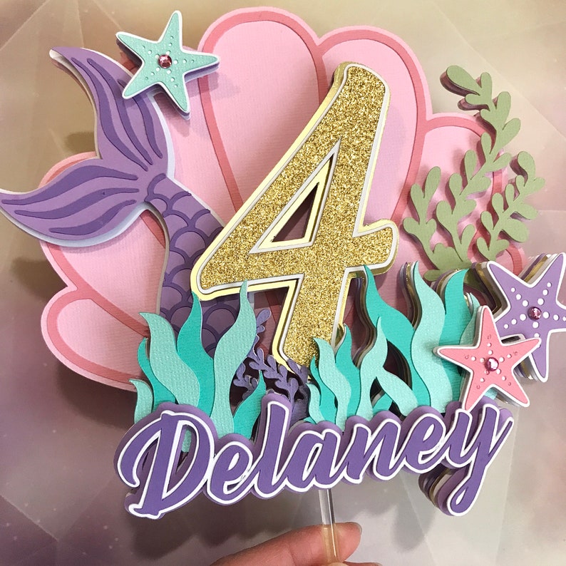 May include: A purple mermaid tail, pink seashells, and green seaweed surround a gold glitter number four and the name "Delaney".