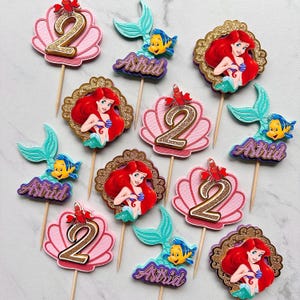 May include: A collection of cupcake toppers featuring characters and motifs from a mermaid theme. The set includes pink seashell cutouts with gold glitter number 2s, teal mermaid tails with yellow fish, and scalloped frames with red-haired mermaids.
