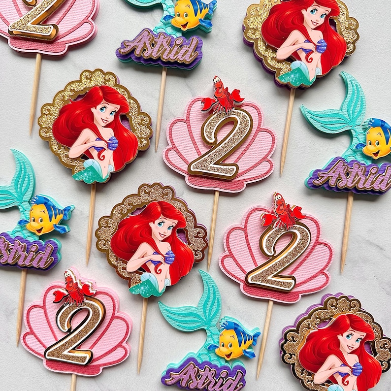 May include: A collection of cake toppers featuring Ariel the mermaid, Flounder the fish, Sebastian the crab, and the number 2, all mounted on wooden sticks. Some toppers include the name Astrid in purple glitter script on a layered background.
