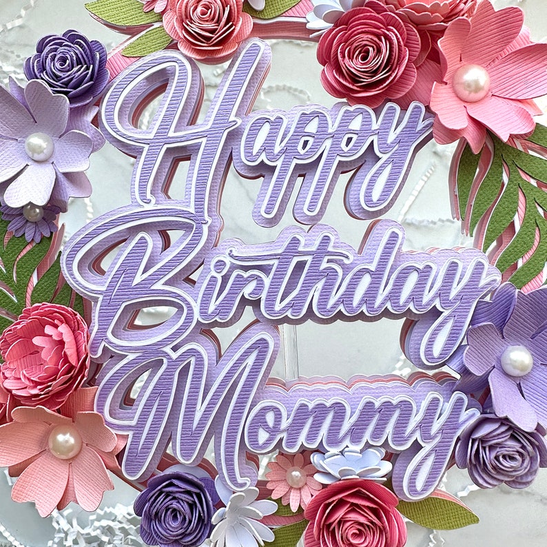 Op de afbeelding: Paarse papieren bloemen caketopper met de tekst "Happy Birthday Mommy" in een gelaagde, 3D-stijl. De topper is versierd met roze en paarse papieren bloemen.
