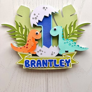 Pu&ograve; includere: Un topper per torta di dinosauro verde, blu, arancione e bianco con il nome "BRANTLEY" su uno stendardo. Il topper presenta un numero uno, due dinosauri e una foglia di palma.