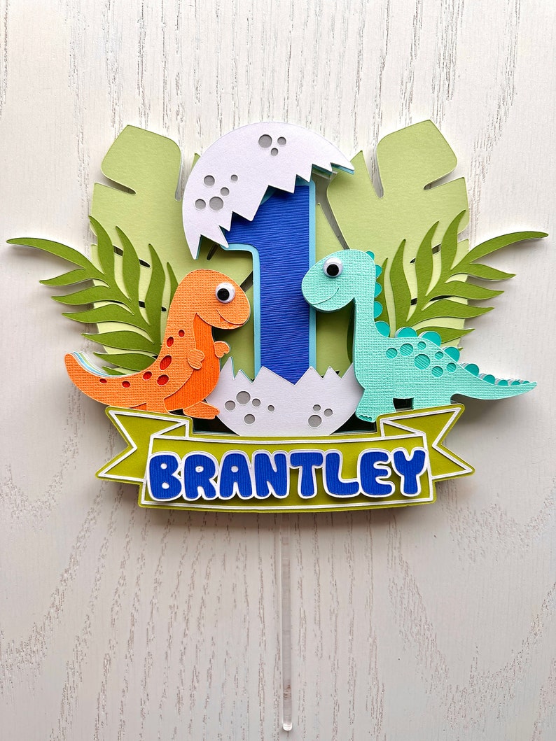 Pu&ograve; includere: Un topper per torta di dinosauro verde, blu, arancione e bianco con il nome "BRANTLEY" su uno stendardo. Il topper presenta un numero uno, due dinosauri e una foglia di palma.