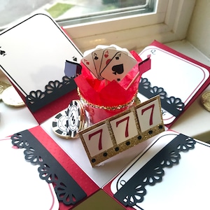 May include: A red and black explosion box with a white background. The box is decorated with black lace and gold coins. The center of the box features a red and gold container with playing cards inside. The container is labeled with the number "777".