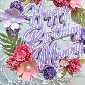 Op de afbeelding: Paarse en roze papieren bloemen en groen omringen een paarse taarttopper met de tekst "Happy Birthday Mommy".