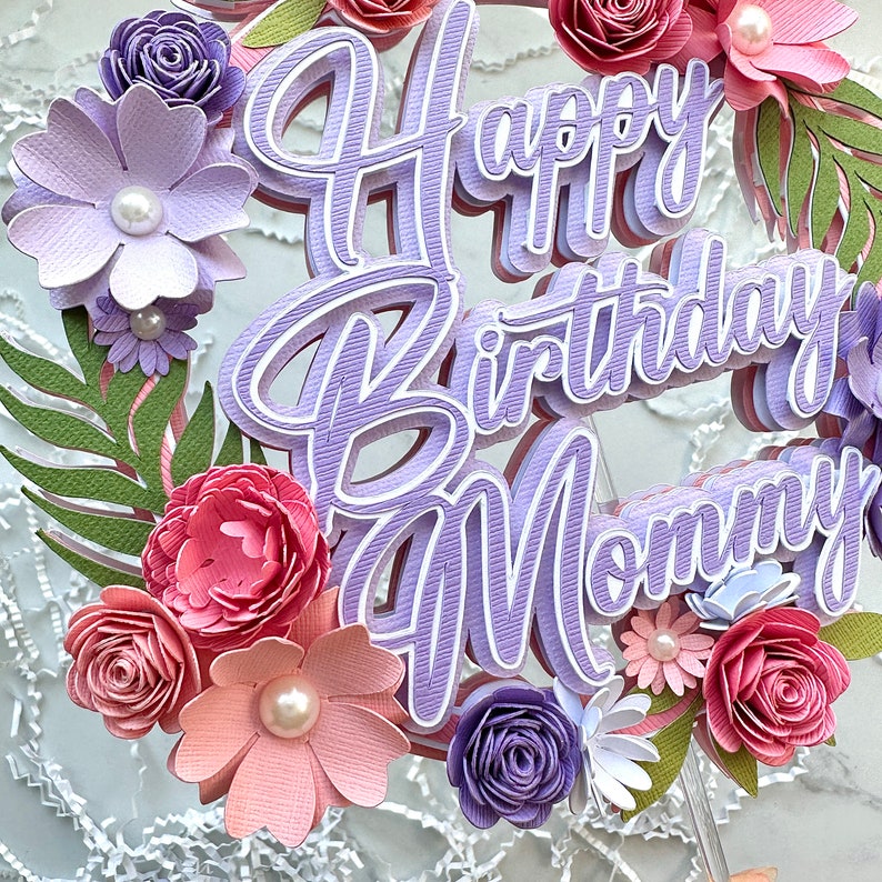 Op de afbeelding: Paarse en roze papieren bloemen en groen omringen een paarse taarttopper met de tekst "Happy Birthday Mommy".