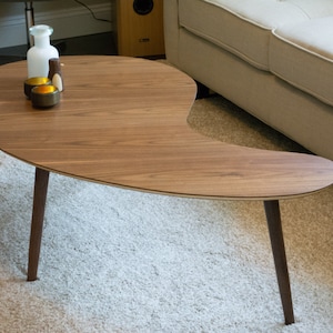 May include: A unique, asymmetrical coffee table with a walnut wood top and dark brown legs. The table has a curved, organic shape and is sitting on a light gray rug.
