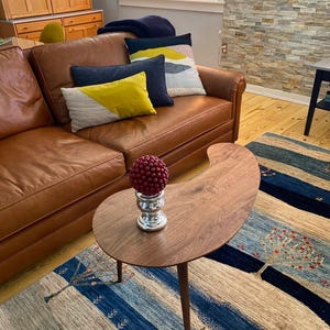 May include: A kidney-shaped wooden coffee table with a smooth brown finish and three tapered legs. On the table sits a silver-toned pedestal base topped with a round, textured, dark red decorative sphere. The table is placed on a patterned rug.