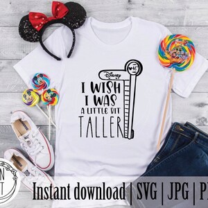 May include: White t-shirt with black text that reads "Disney I wish I was a little bit taller" with a black and white illustration of a height chart next to it. The shirt is laying on a wooden surface with a pair of white sneakers, a red and black headband, and a colorful lollipop.