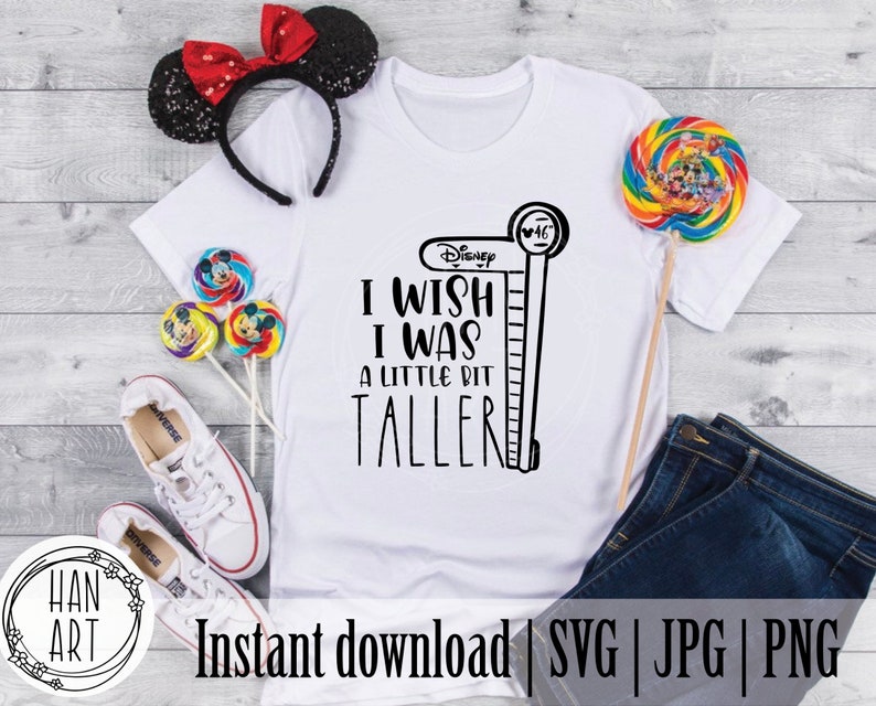 May include: White t-shirt with black text that reads "Disney I wish I was a little bit taller" with a black and white illustration of a height chart next to it. The shirt is laying on a wooden surface with a pair of white sneakers, a red and black headband, and a colorful lollipop.