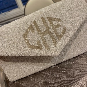 May include: A white beaded clutch bag with a silver monogram of the letters "CRE".
