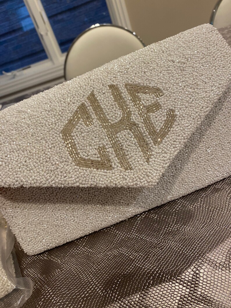 May include: A white beaded clutch bag with a silver monogram of the letters "CRE".