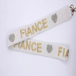 May include: White beaded bag strap with gold lettering that says "FIANCE" and silver heart accents.