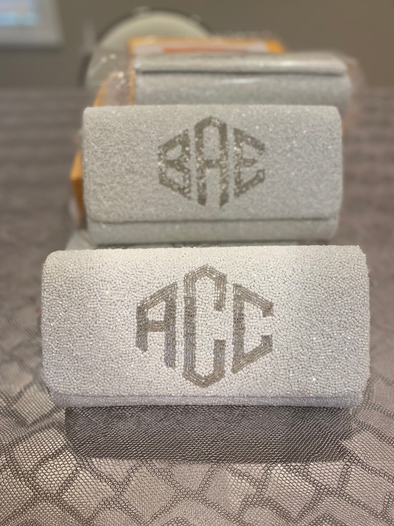 May include: Two white beaded clutches with silver monogrammed initials. The top clutch has the initials "BAE" and the bottom clutch has the initials "ACC".