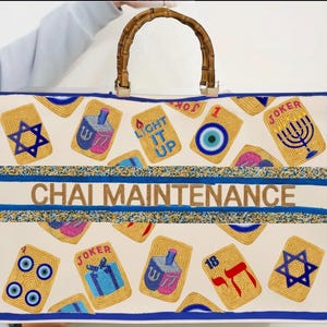 May include: A rectangular canvas tote bag with a bamboo handle, featuring a cream background with a pattern of embroidered mahjong-style tiles including Stars of David, dreidels, and evil eyes. The text CHAI MAINTENANCE is centred.