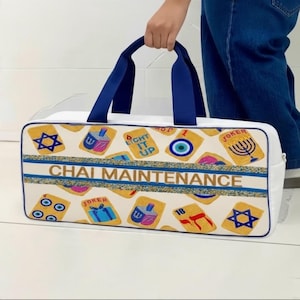 May include: A white rectangular duffel bag with dark blue handles and trim. The bag features a repeating pattern of gold-toned cards with Jewish symbols and the text "CHAI MAINTENANCE" printed in gold across a central horizontal blue stripe.