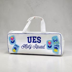 May include: A white rectangular zippered bag with two top handles, featuring blue embroidered text that reads UES Mahj Squad. The bag is decorated with four beaded patches depicting colorful flowers and playing card motifs in blue, pink, and yellow.