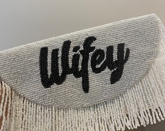 Può includere: Clutch bianca con perline con una scritta "Wifey" in perline nere e frange bianche.