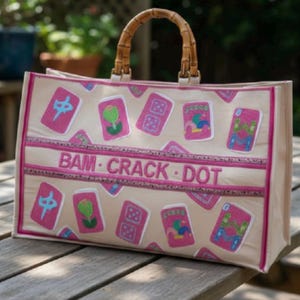 May include: A beige tote bag with bamboo handles featuring a pattern of pink Mahjong tiles and a central horizontal band with the text BAM CRACK DOT in pink lettering. The bag has pink trim along the edges and a structured rectangular shape.