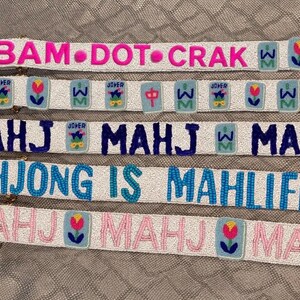 May include: Five white beaded straps featuring colorful embroidered patches and text. The straps display phrases including BAM DOT CRAK, MAHJ, and MAHJONG IS MAHLIFE in pink, dark blue, light blue, and light pink beads, accented by floral and character icons.