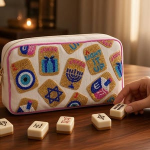 May include: A rectangular white canvas pouch with pink piping, decorated with colourful beaded patches featuring a menorah, a dreidel, a Star of David, an evil eye, and the text LIGHT IT UP. Several mahjong tiles are scattered in front.
