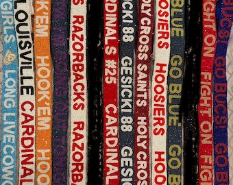 May include: A collection of colorful beaded straps with various team logos and slogans, including "Go Bucs!", "Fight On", "Go Blue", "Hoosiers", "Holy Cross Saints", "Gesicki 88", "Cardinals #25", "Razorbacks", "Hook 'Em", "Louisville Cardinals", and "Live Cowgirls".